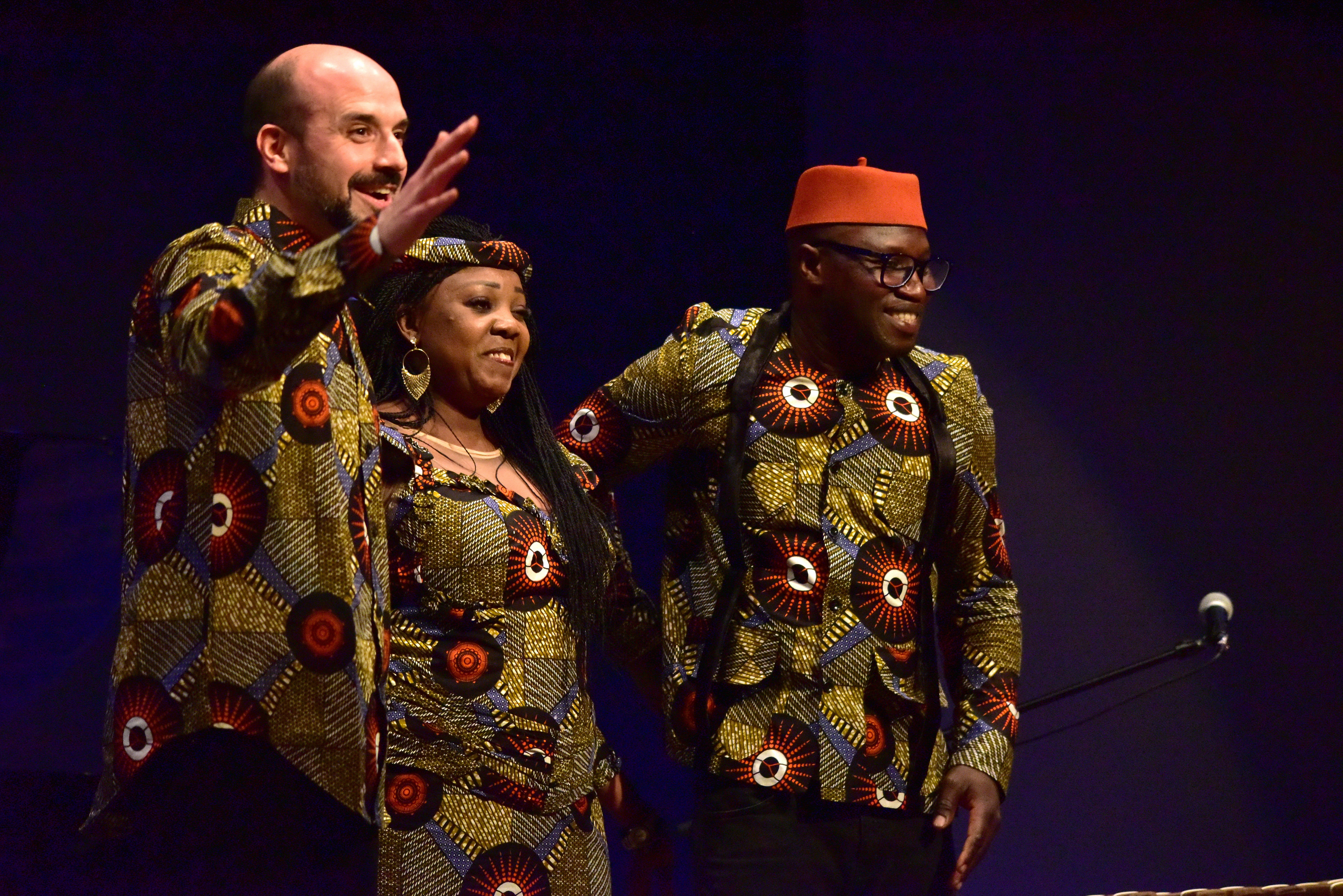 Claude Diallo greeting the audience on stage, with Aminata Khass and Djiby Diabate beside him in matching West African dress