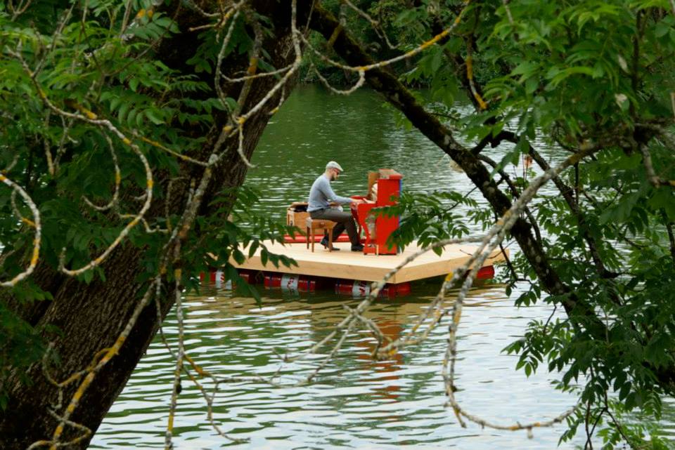 Claude Diallo playing a red upright piano on a wooden pontoon floating on a lake, seen through overhanging trees